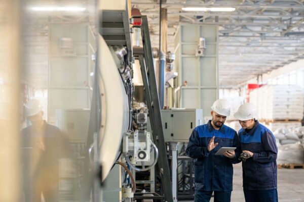 Two engineers in hard hats reviewing a tablet on a manufacturing plant floor