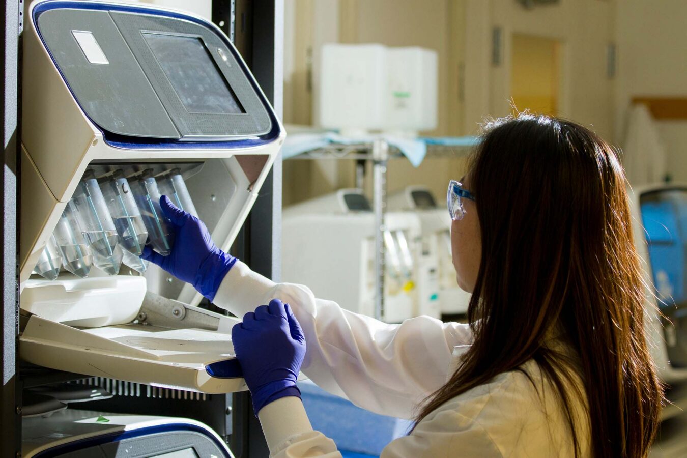 Engineer in a lab coat and safety glasses handling sample tubes in a laboratory testing instrument